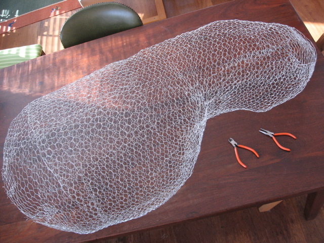 A 3D armature made of bird-netting in the shape of a large peanut. It is lying on a wooden table with two small pliers.