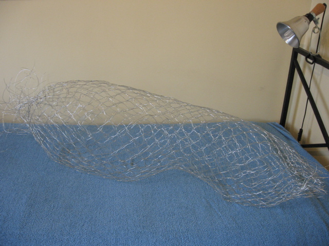 A 3D wire hollow lace form lying on a blue bedspread on a bed. There is a tangle of wire ends sticking out of one end of the lace.