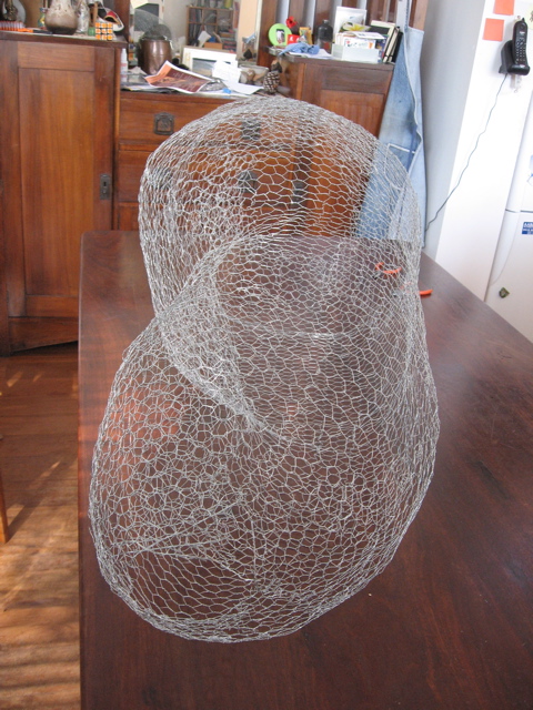 The bird-netting armature sitting up on a wooden table, with a wooden dresser in the back ground