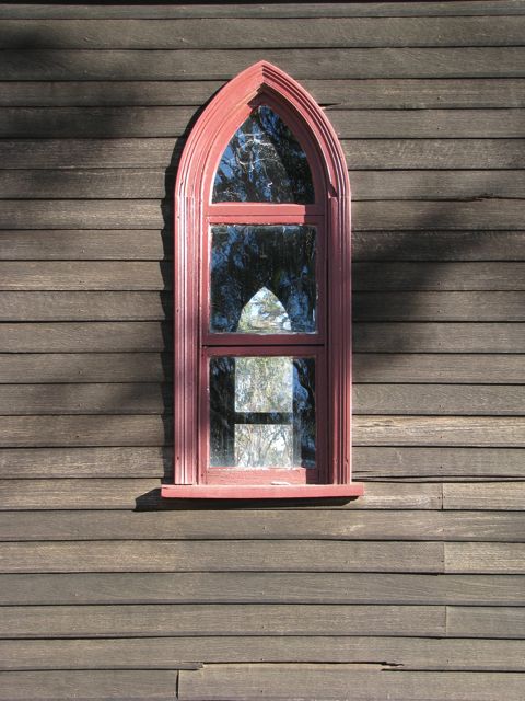 A church wall of unpainted, weathered grey jarrah weather boards, with a red-painted gothic-shaped window in the centre. Through the window can be seen the matching window on the opposite side of the church, looking out onto gum trees. There are tree shadows cast on the wall.