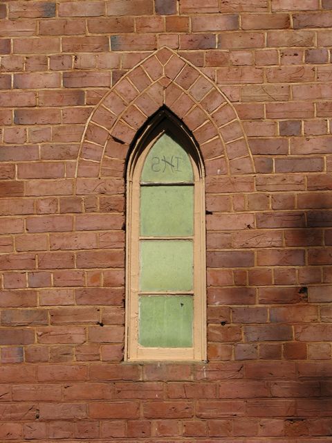 An old orange brick church wall with a narrow gothic-pointed window. The glass is a light green and in the top pointed panel of the window are the letters IHS, reversed. There are tree shadows cast on the bricks.