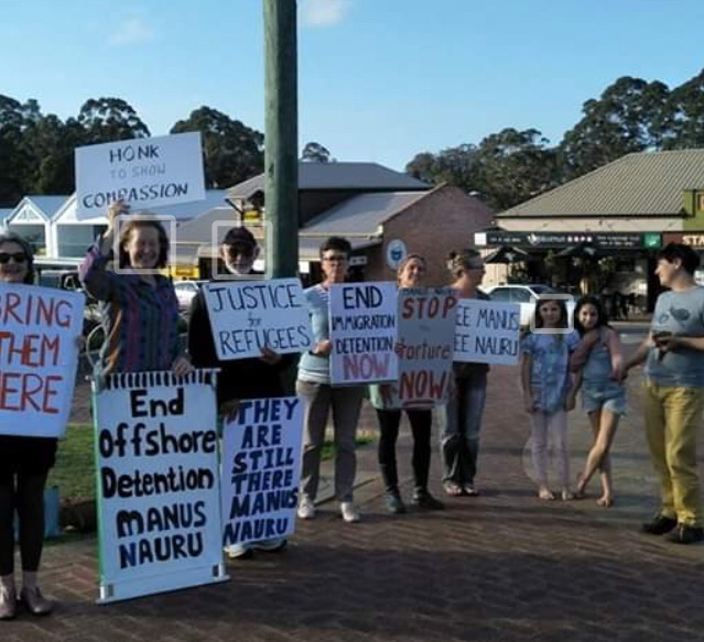 A group of 6 people holding placards protesting for refugee freedom, standing on a street corner. A women and two girls stand talking to the protestors.
