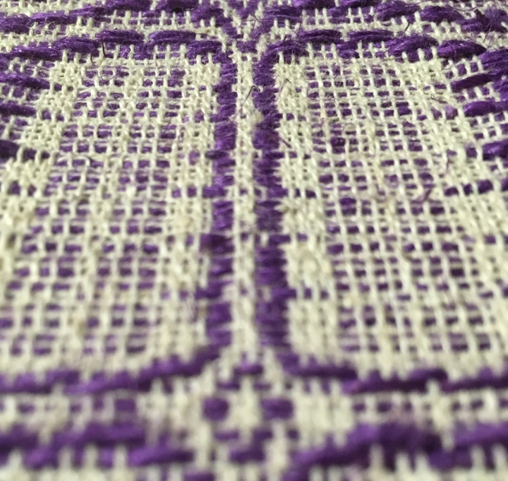 Closeup of handwoven cloth. Thick purple threads weave through thinner undyed threads. The pattern is geometric, the photo is partly out of focus.