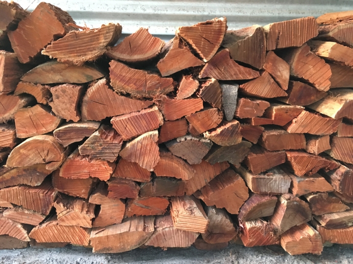 Load of cut & split red-brown wood is stacked neatly against a corrugated metal wall.