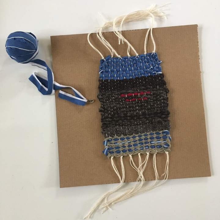 A small weaving and ball of fabric strips lies on a square of cardboard